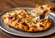 Hot specialty pizza on a round metal pan with a slice being lifted, featuring melted mozzarella, savory meat toppings, and stretchy cheese.