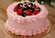 Round pink frosted cake decorated with piped buttercream, fresh strawberries, blackberries, and white candy pearls on a gold cake board.