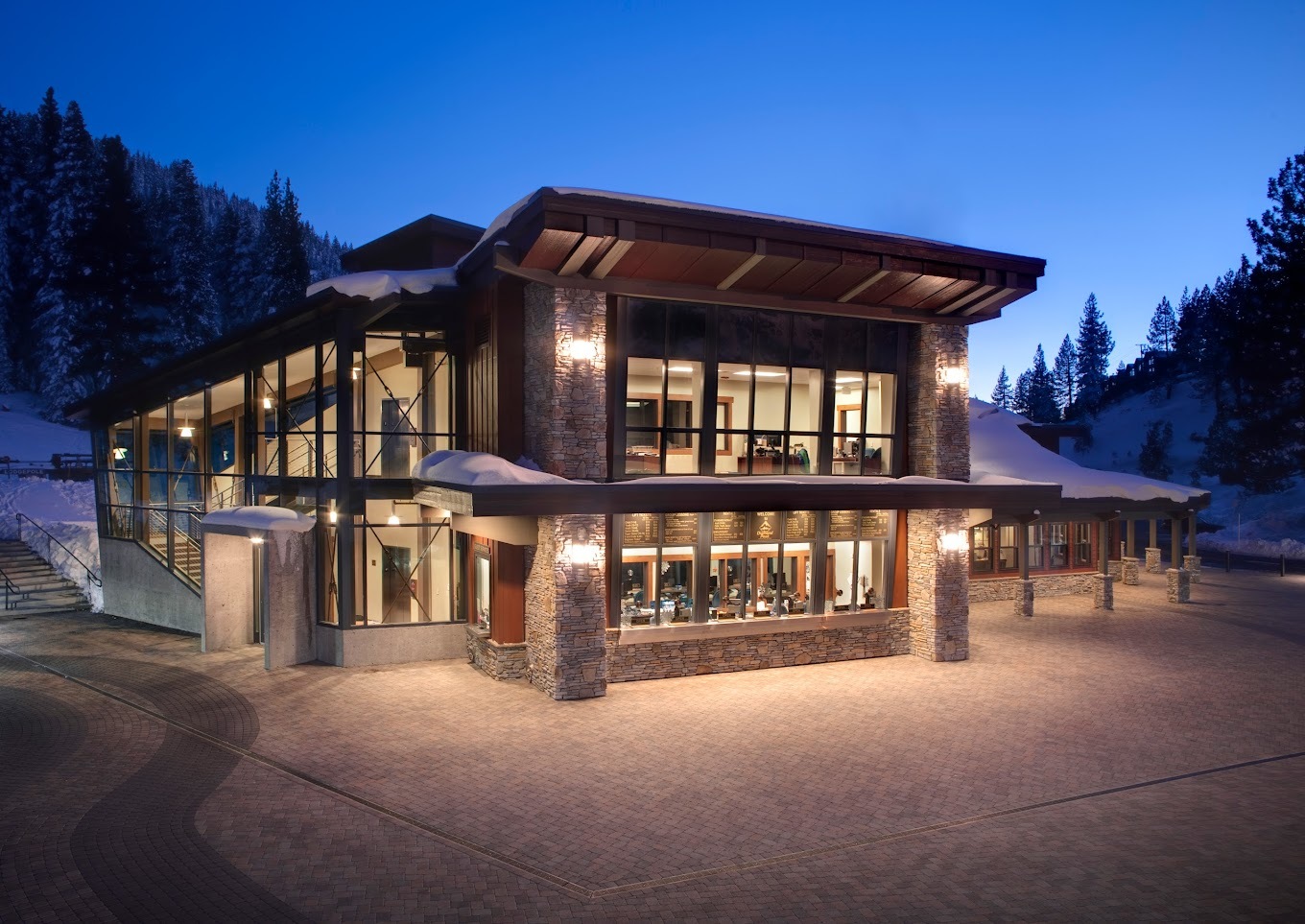 A large brown building surrounded by trees and hills, large windows display warm light onto the concrete around the building.