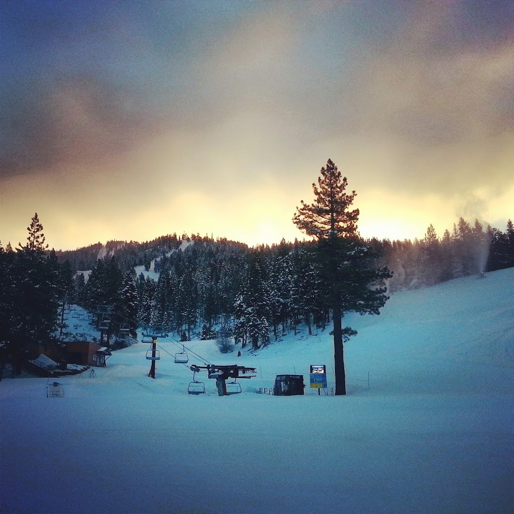 A snowy hill covered in pine trees and snow, with a beautiful golden sunrise in the sky. On the hill the lift for skiers.