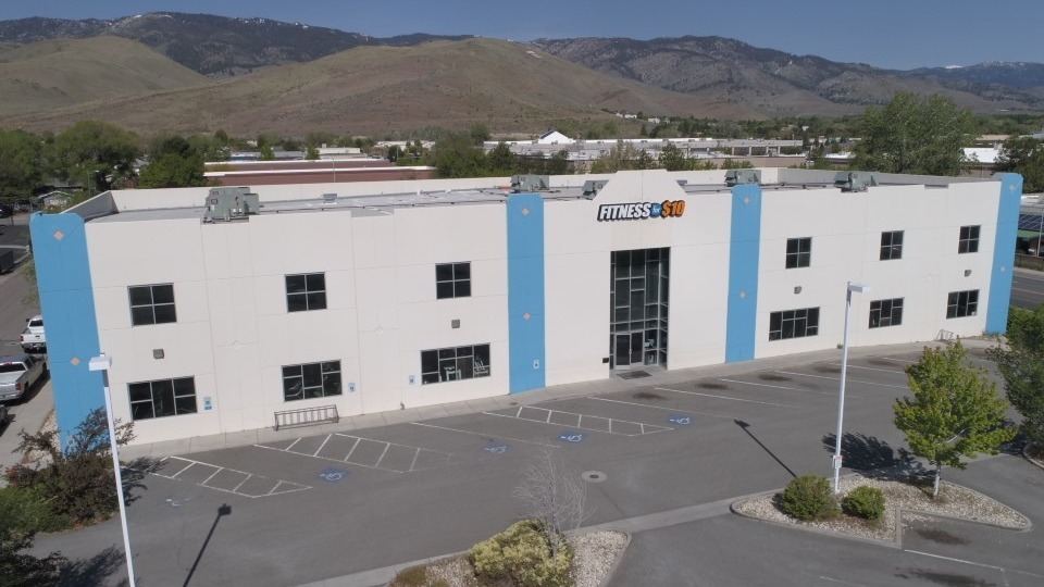very large white building with blue accenting on the corners and by the door. A sign above the door and windows in the center of the building reads "Fitness for 10"