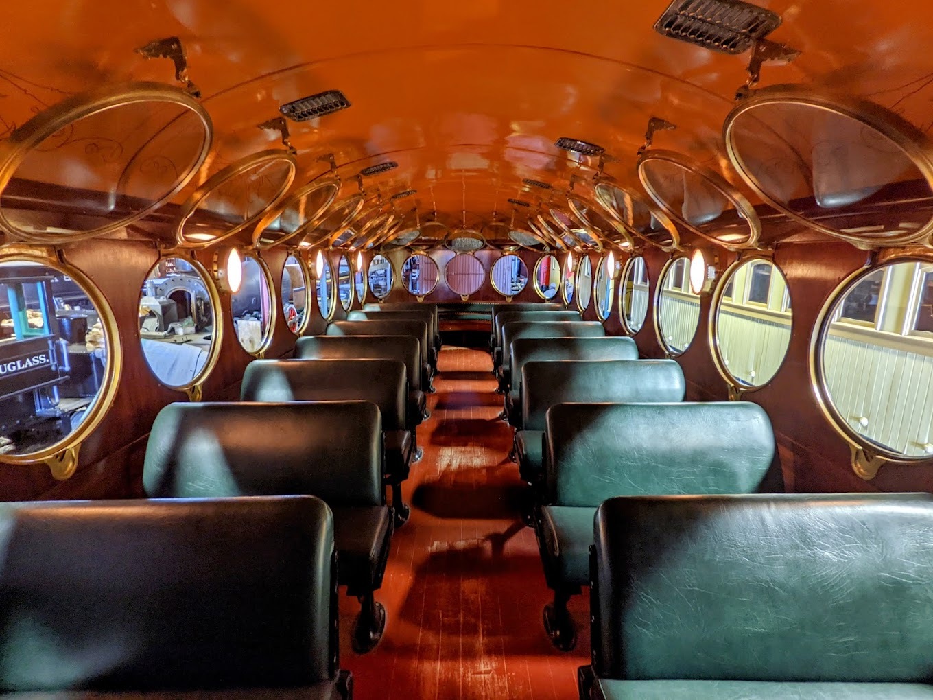 An inside of a people moving train, the windows are circular and every window is open. The aisle has red carpet and each row of seats is a green leather bench.