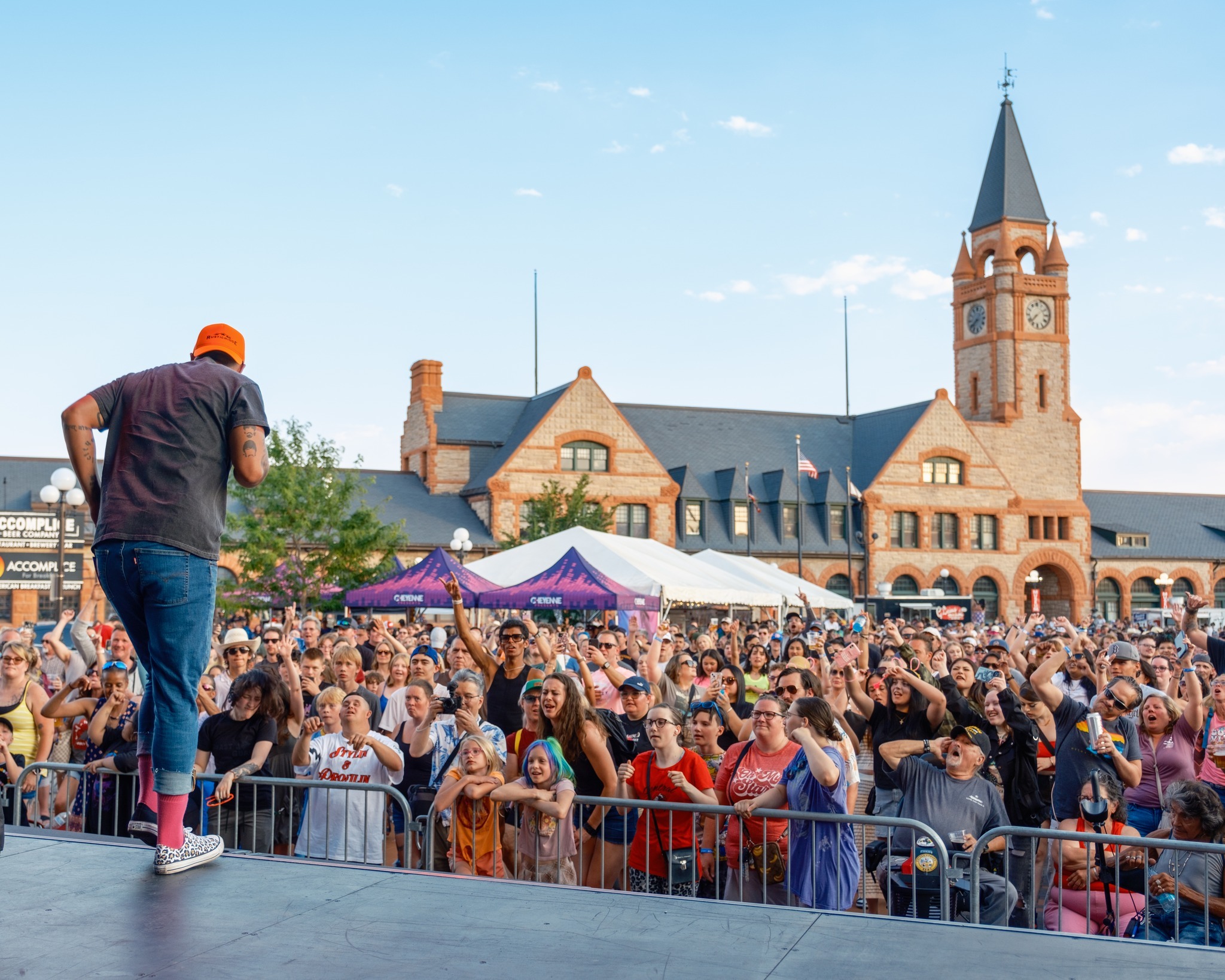 A view of a performer singing on stage to a large crowd of people in a plaza with large historical buildings in the background.