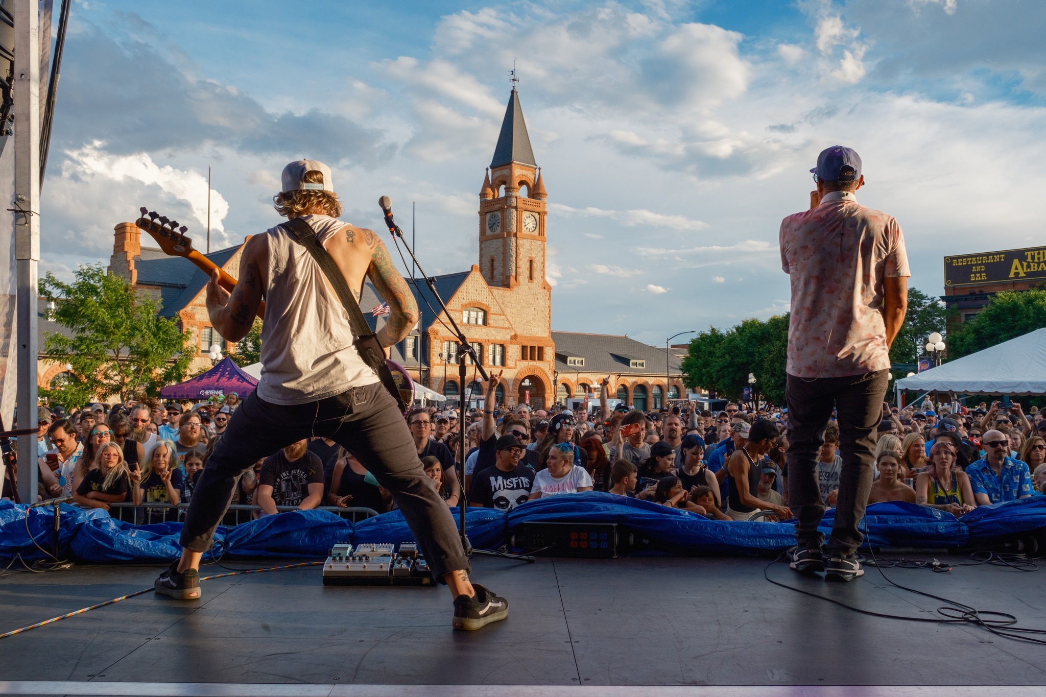 A view of two performers on a stage, signing and playing guitar in a plaza with a crowd full of people.