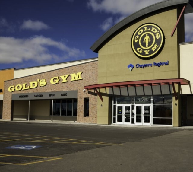 An outdoor view of a gym with yellow lettering above the windows of the building and an awning over the glass entrance doors.