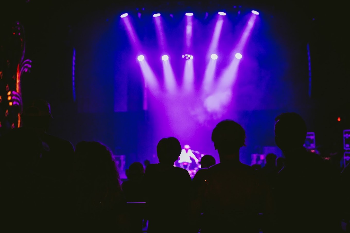 A view of a stage from the crowd, a band performing on stage with lights illuminating purple.