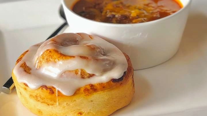 A view of a cinnamon roll with a white cup full of chili.