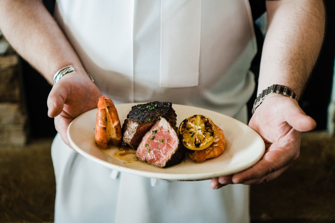 A chef displaying a dish on a white plate- the plate consists of a cooked steak along with an lemon.