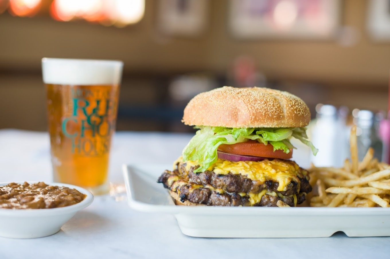 A white plate with a burger, and a side of fries alongside, to the left there is a white bowl of chili and a large glass of beer.