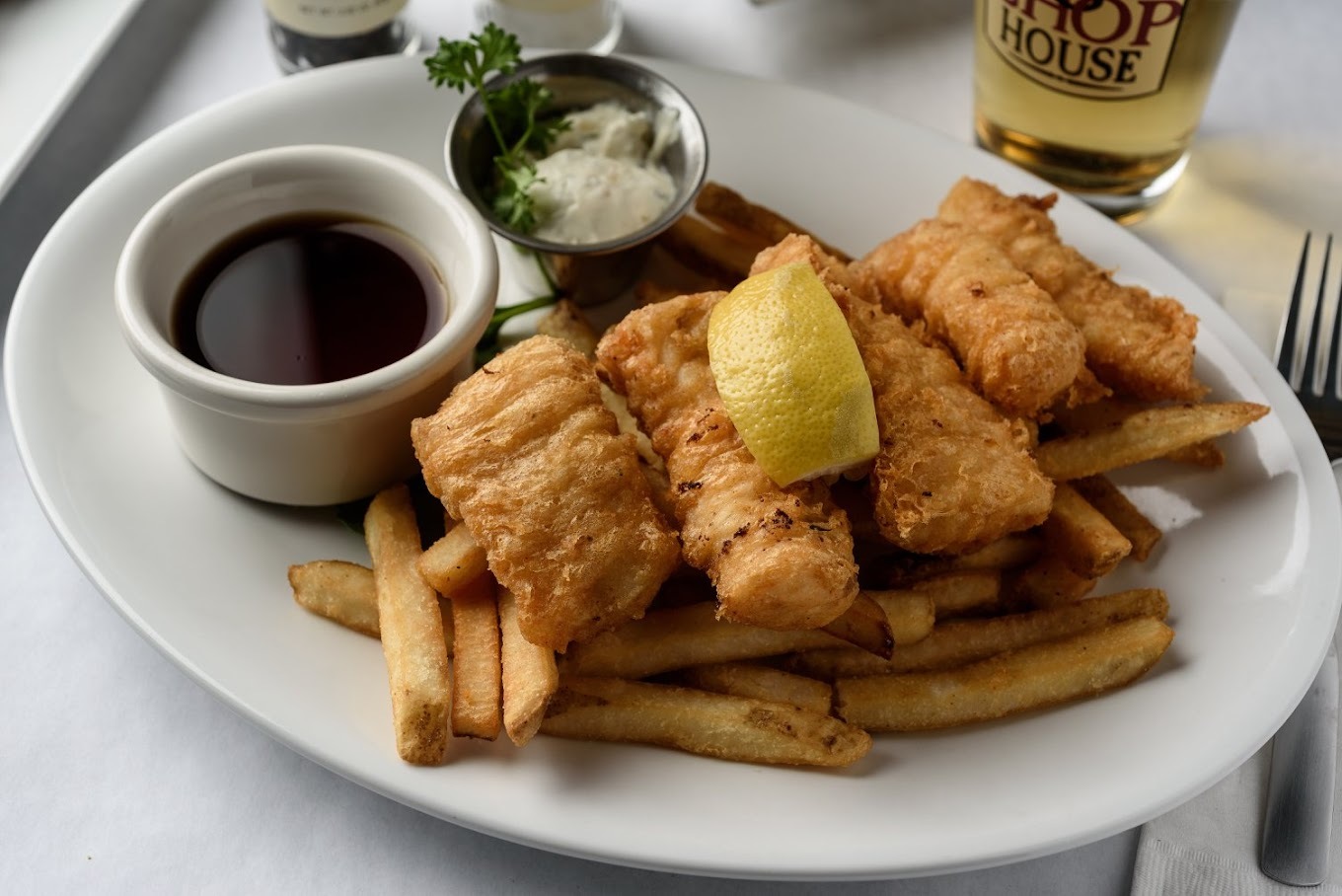 A white plate with French fries, and chicken strips placed on top of the fries with a lemon slice. Served with a side of sauce and butter.