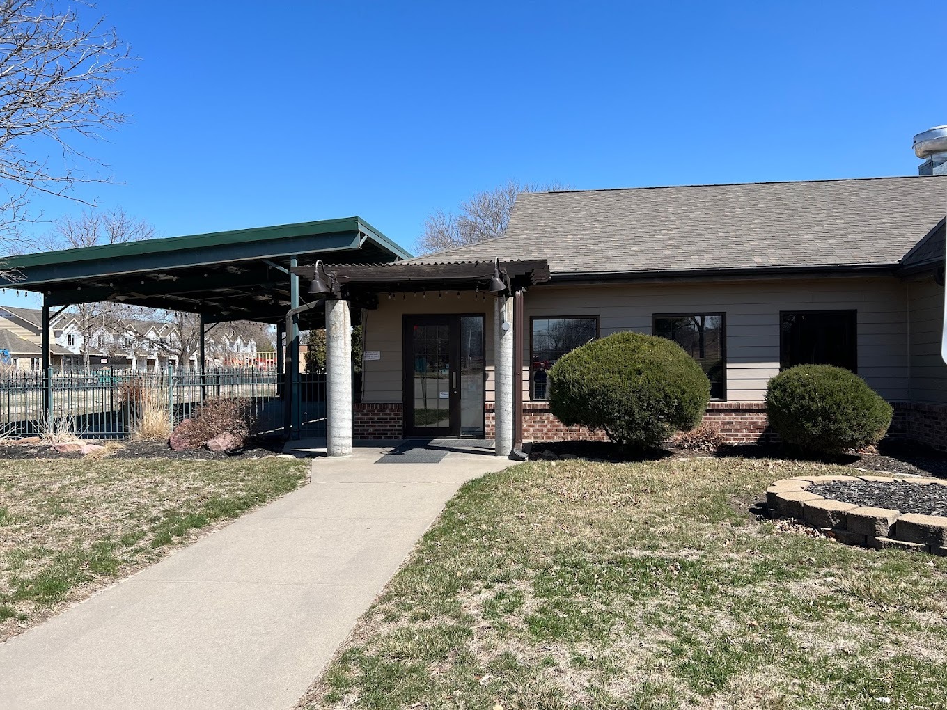 An outside view of a building with a large awning out front, in front of a large grassy area with a sidewalk leading up to the building.