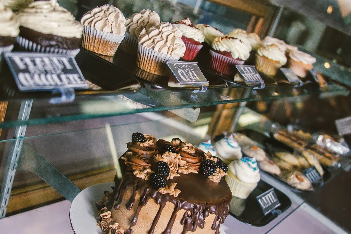 A view of baked goods on top of a glass shelf, a variety of cupcakes, cake, and cookies.