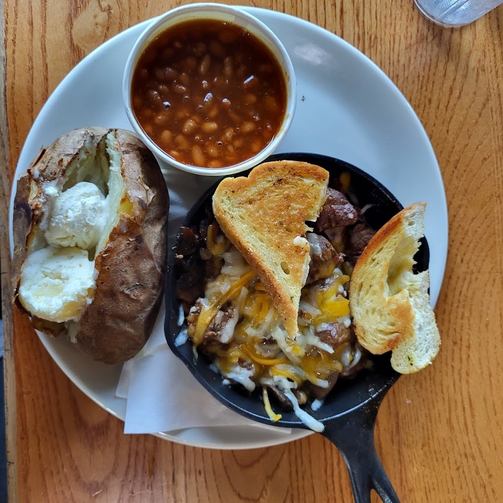 A white plate with a baked potato, a white bowl of beans, and a black skillet with meat and toast on a wooden table.