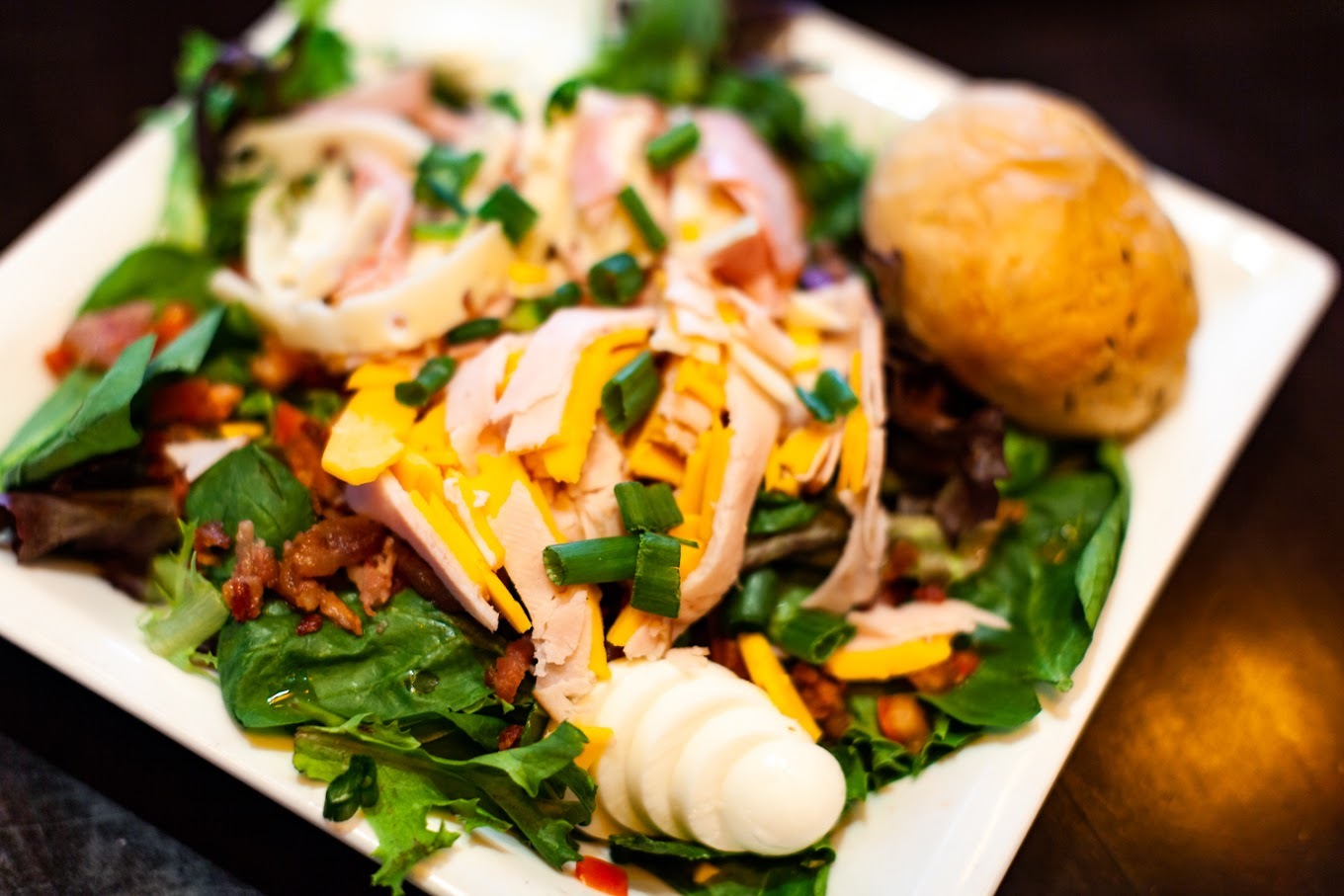 A white plate consisting of a chef salad topped with meat, hard boiled eggs, cheese and a bread roll on the side.