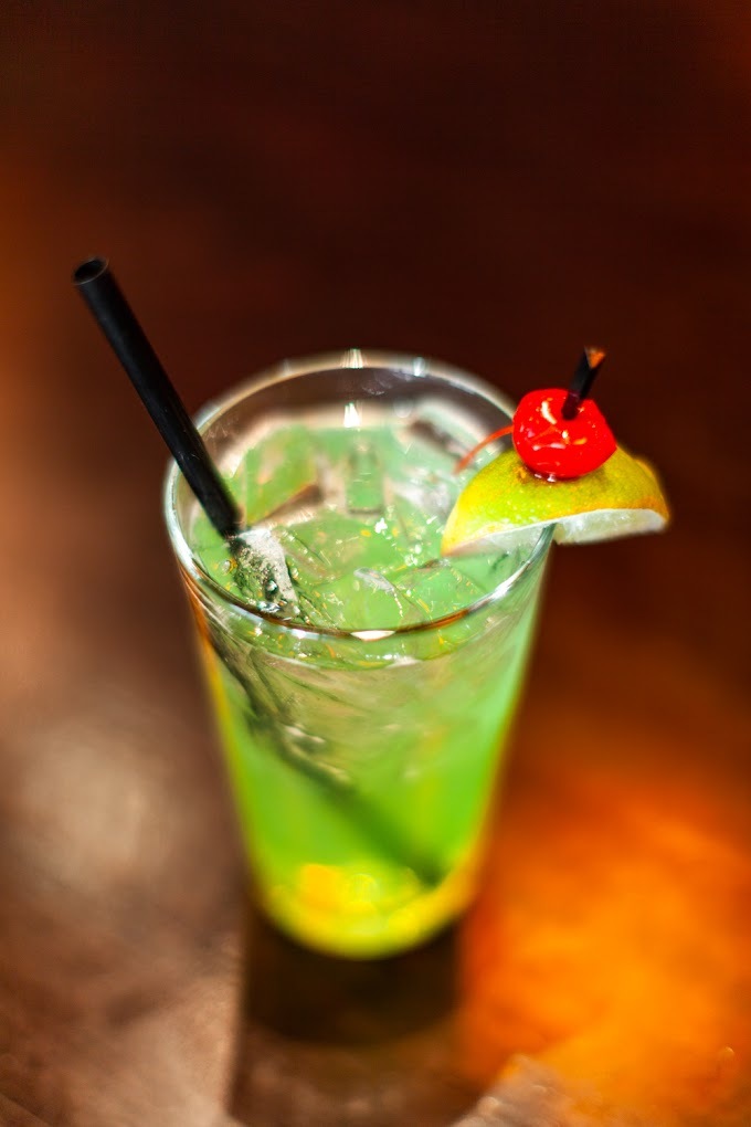 A large mixed drink that is green and topped with ice and a garish on the side of a lime and a cherry.