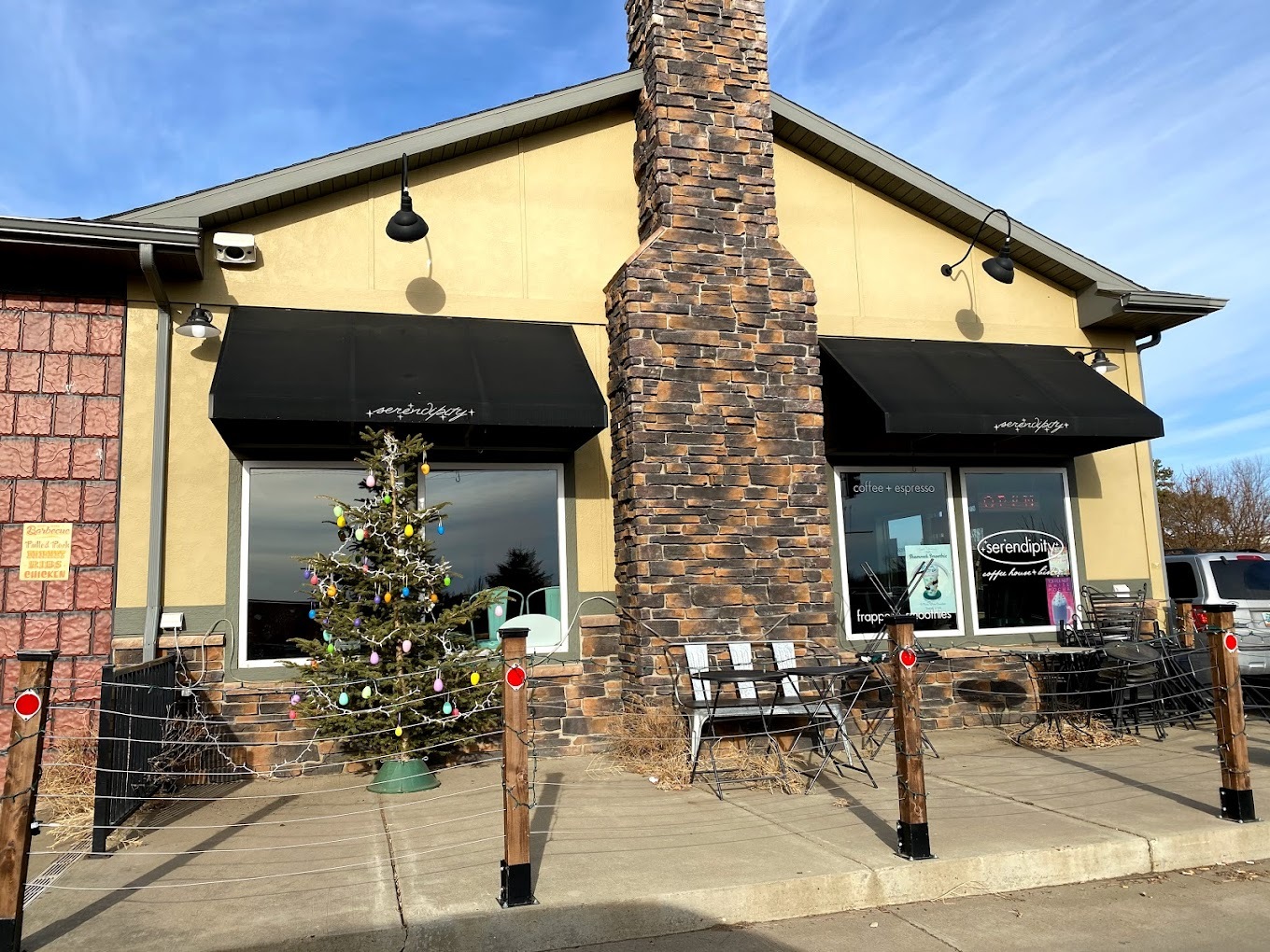 An outside view of a cafe, a small yellow building with brick accenting, lighting trees out front, and a bench.