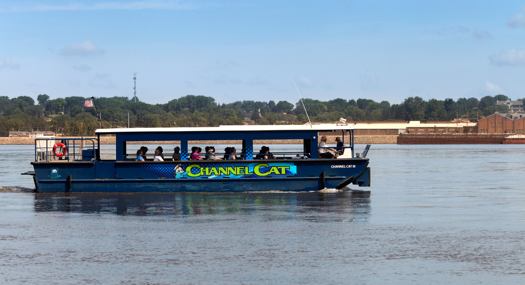 Blue boat with the words "Channel Cat" written in green on it and passengers inside the boat. This boat is in the water.