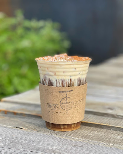 Small iced coffee with a brown cup holder that has the words "Iron + Grain Coffee Company" stamped on it. The background is a wooden table and green shubbery.