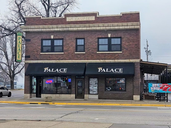 Exterior view of a brick building with sun shade, seating, and a black fence on the right side. The sunshade above the windows have the words "Palace" written on it. The very edge of the building has a green and yellow sign that also says "Palace."