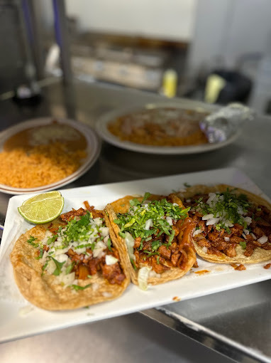 Three tacos on a rectangular white plate. Two plated dishes are in the background.