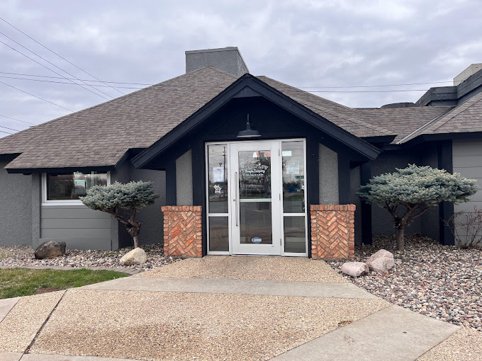 Exterior view of a house like gray building. Either side of the entrance has red brick columns, small trees, and rock landscaping.