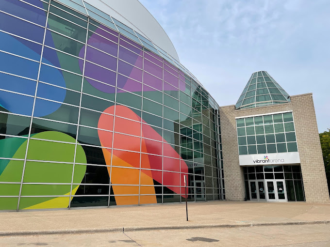 Upward view of the entrance to a beige and glass building. The left side of the image shows a colorful pattern within the window panes.
