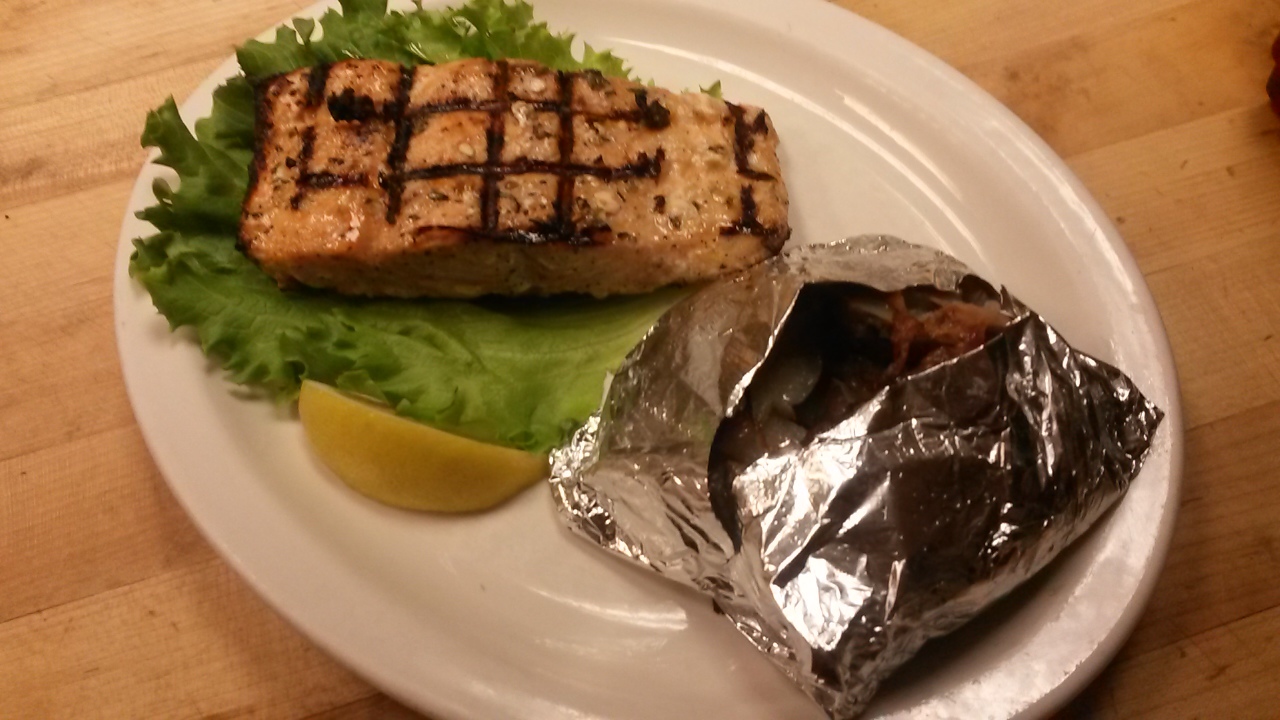 Grilled chicken on a piece of lettuce next to a lemon slice. Off to the side is a baked potato in foil.