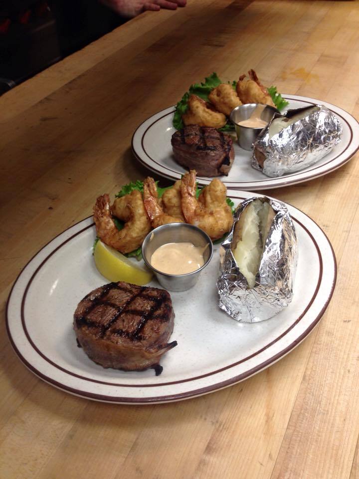 Two plates containing steak, shrimp, lemon slices, cups of sauce, and foiled baked potatoes.