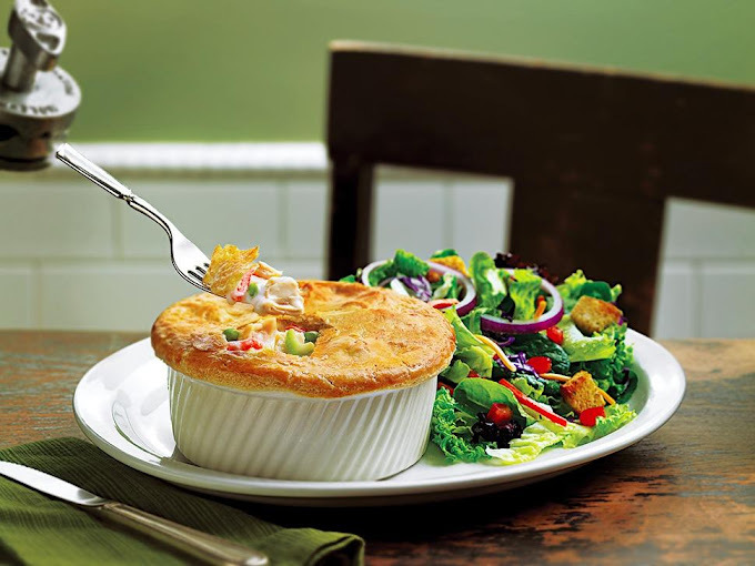 Baked dish in a white striped bowl. A fork is scooping a piece out. Next to it is a salad with croutons and onions. In the background there is a chair, white tile, and green walls.