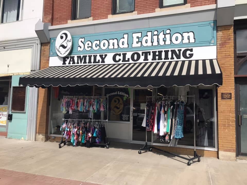 Downtown brick building. There is a black and white striped sun canopy. The front entrance has two racks of clothing. The sign above the door is in teal and reads "Second Edition, family clothing."