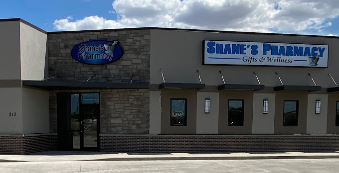 Flat, beige building with numerous small windows. The sign above the door entrance is circular and blue. The sign off to the side and above the windows is large and white. It reads "Shane's Pharmacy" in blue lettering.