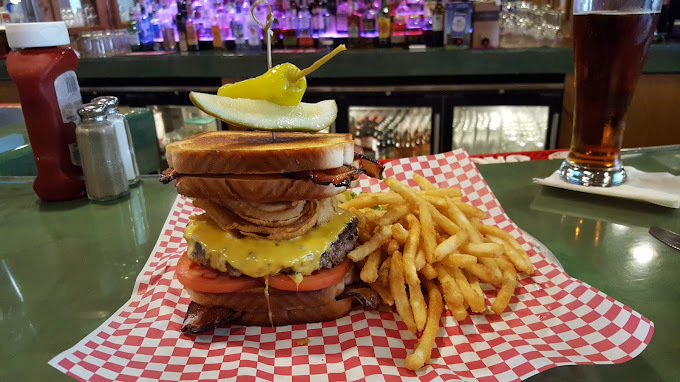 Sandwich with four bread slices, patty, tomatoes, cheese, and pickles, held together by a long toothpick. There is a side of fries, and both items sit on a red and white checkered paper.