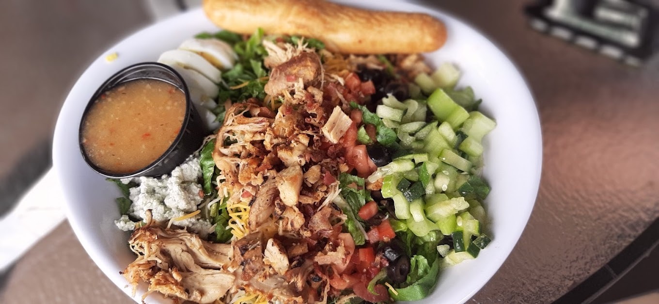 A white bowl consisting of a salad with cucumbers, chicken, egg, feta and shredded cheese. Served with a side of sauce and a breadstick.