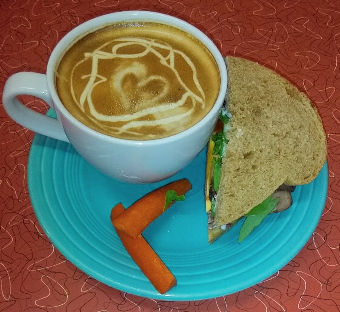 A blue serving plate consisting of a latte with a heart latte art, and a side sandwich and a couple of carrot sticks.