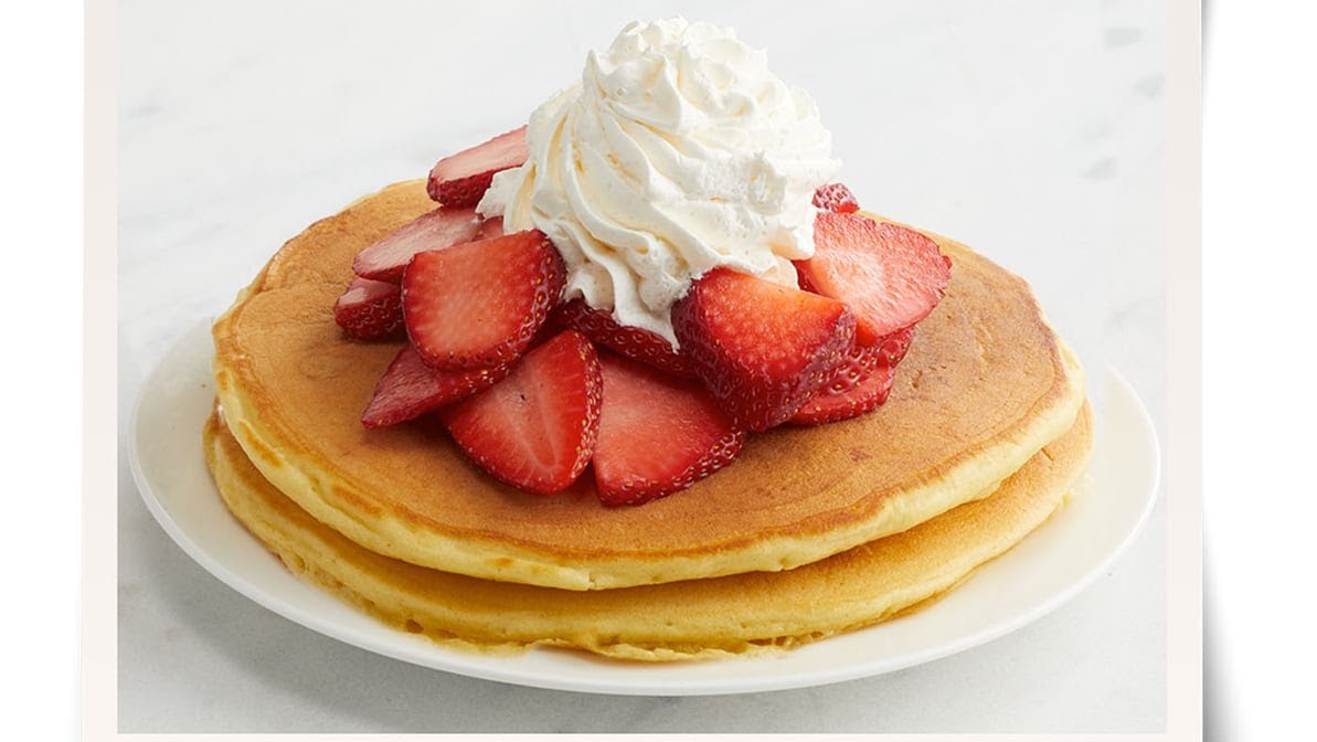 A white plate consisting of a stack of two pancakes, topped with sliced strawberries, and a large dollop of whipped cream.