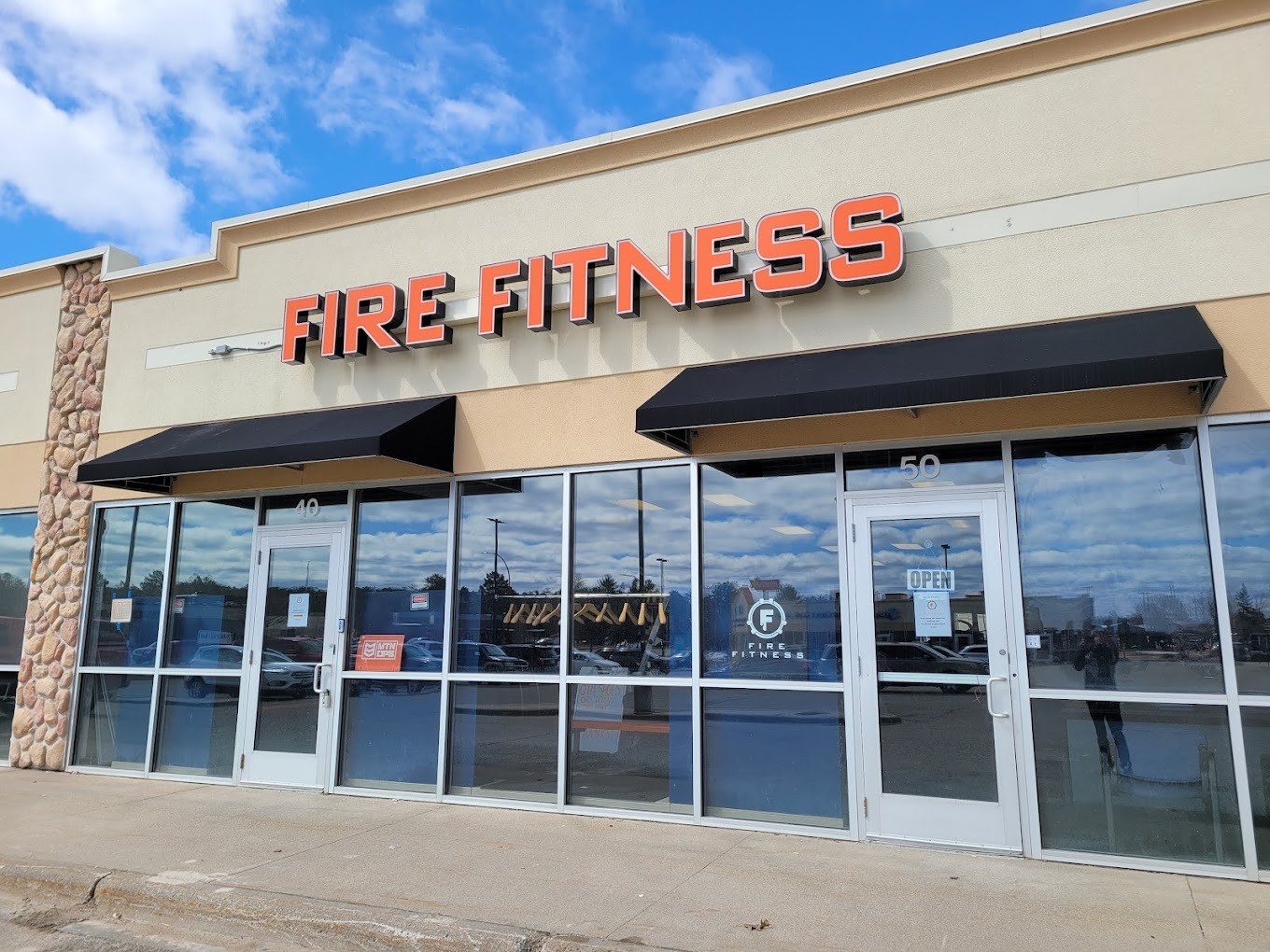 An outside view of a gym inside of a strip mall, windows line the entrance with awnings covering the windows. Lettering in orange reading "Fire Fitness" above the window awnings.