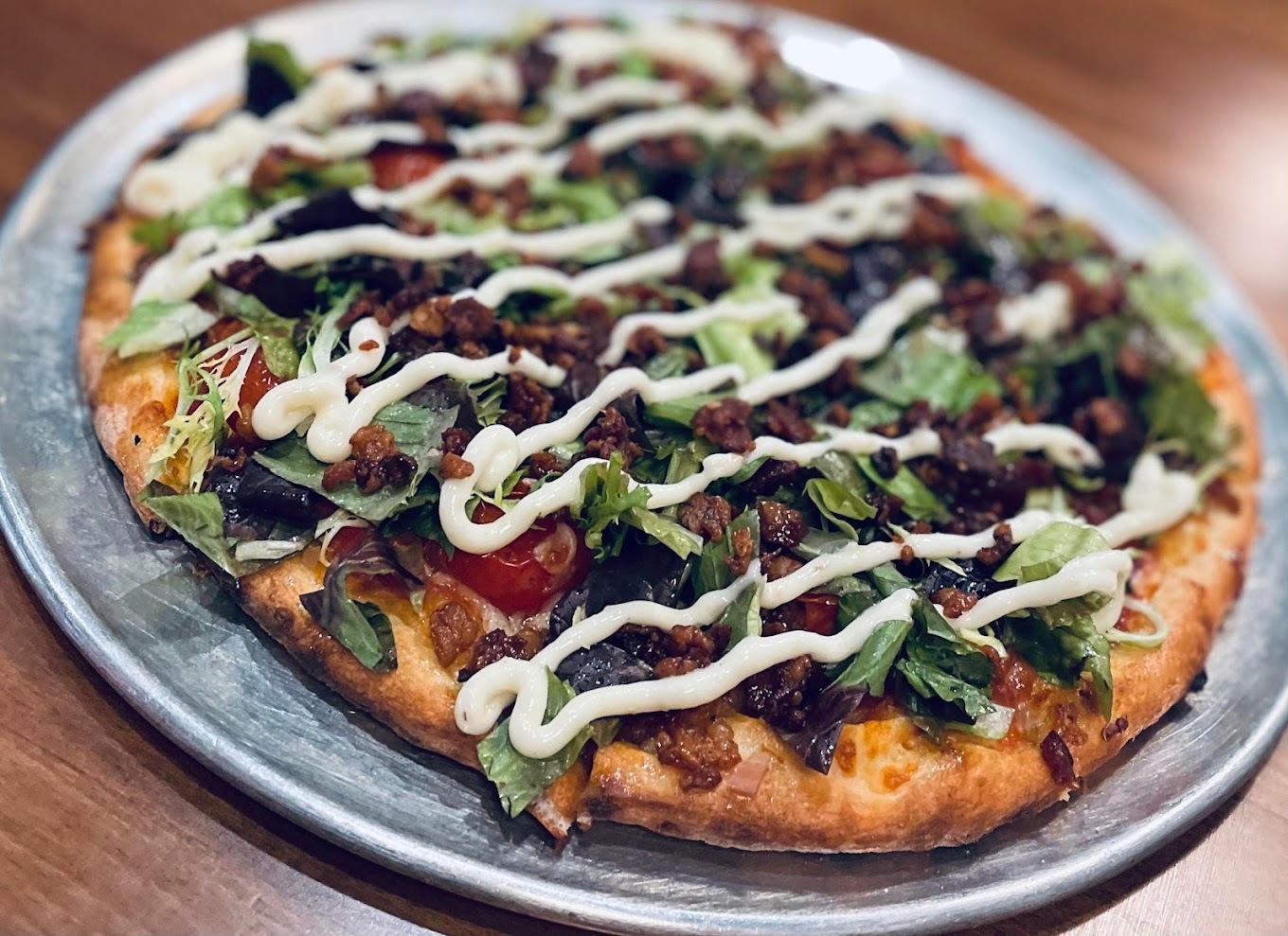 A loaded pizza on a silver serving platter with sauce, cheese tomatoes, leafy greens and drizzled with sour cream.
