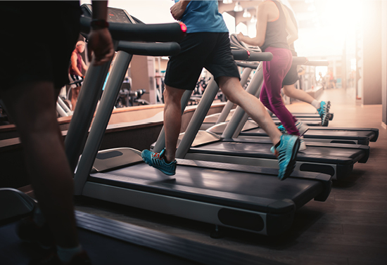 Image of people running on a set of treadmills. The upper halves of their bodies are not visible. The focus is a man in the center with a blue shirt, black shorts, and blue shoes.