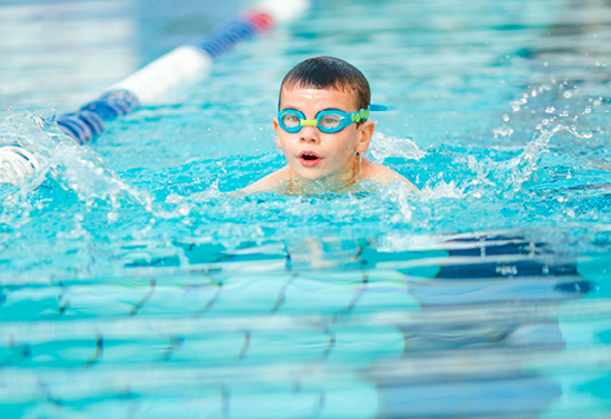 Child with green swim googles swimming in a pool with a floating lane bar on the left.