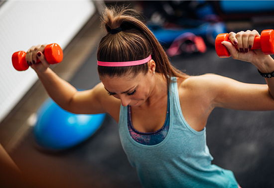 Woman with brown hair, pink headband, and a blue tank top lifts two pink weights.