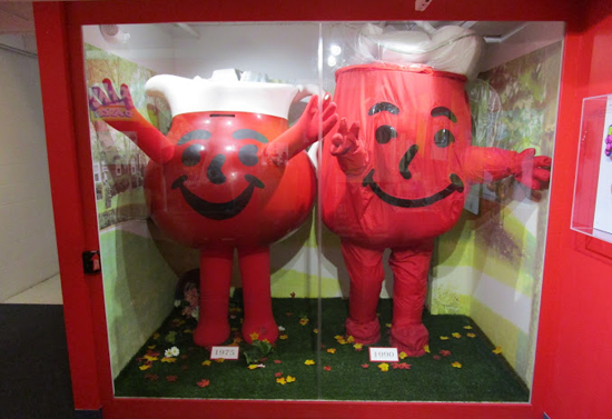 Image of two red kool-aid costumes in a red and glass display case.