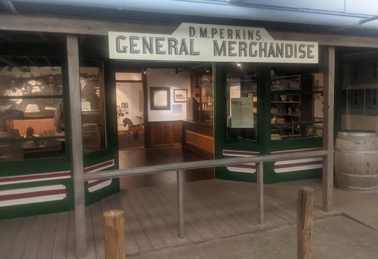 Front view of building entrance, with windows on either side. Sign above door states "D.M. Perkins General Merchandise."
