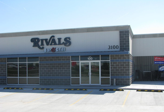 Grey brick building with parking lot in the front. Sign above door and windows states "Rivals Bar and Grill."