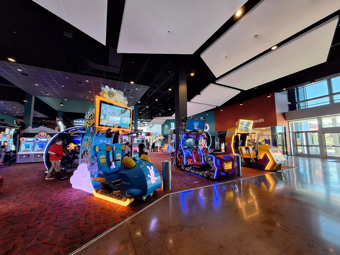 A large room with windows and a door to the right- on the left there is a dimly lit area filled with lit up arcade games.