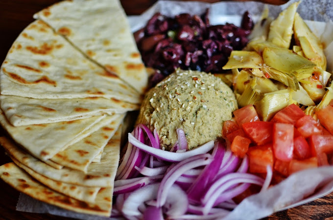 An view of a serving tray filled with tortilla's, cut up onions, tomatoes, hummus, peppers, and olives.