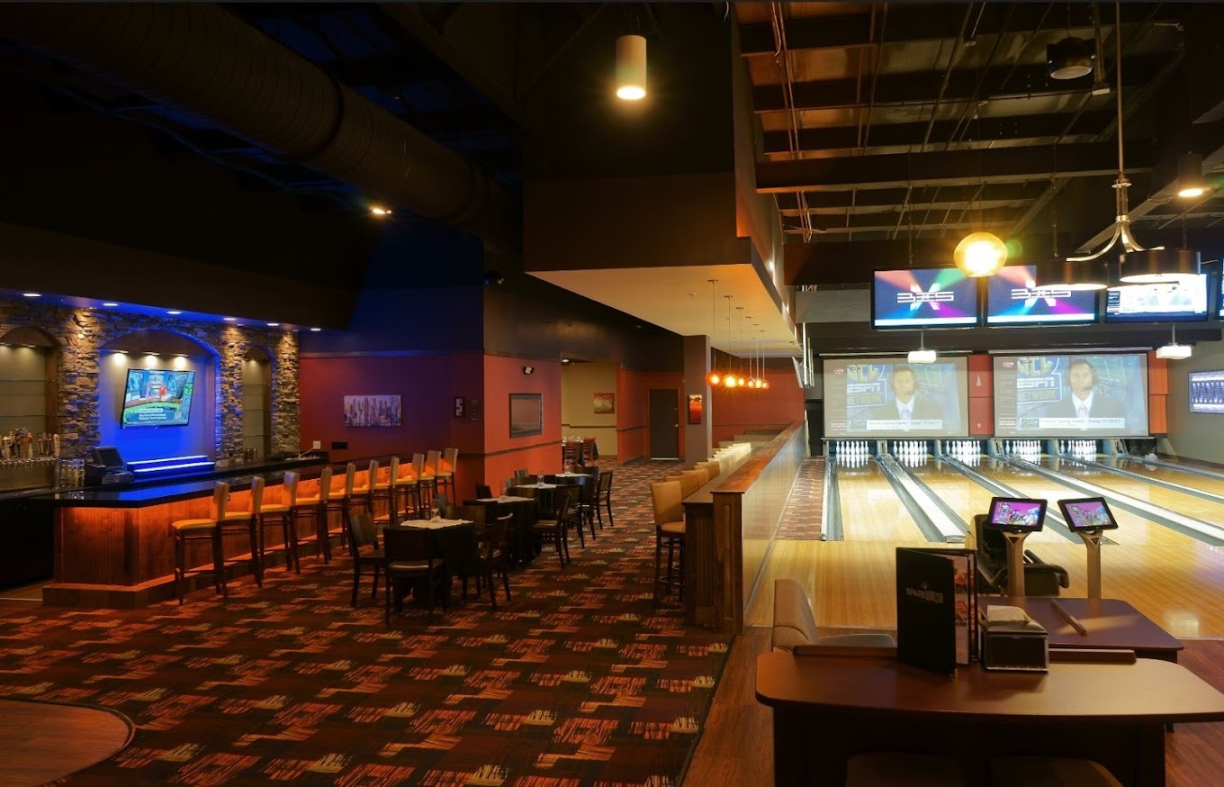 A view of a bowling alley, bowling lanes are to the right in a well light area and off to the left side there is a bar area that is dimly lit.