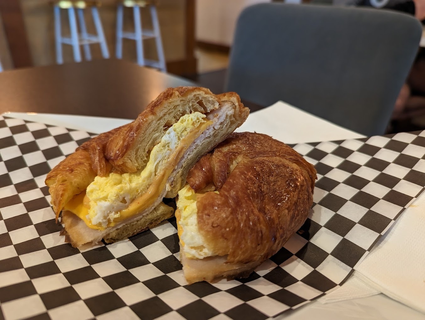 A view of a sandwich with a checkered paper, a croissant breakfast sandwich consisting of egg, cheese and meat.