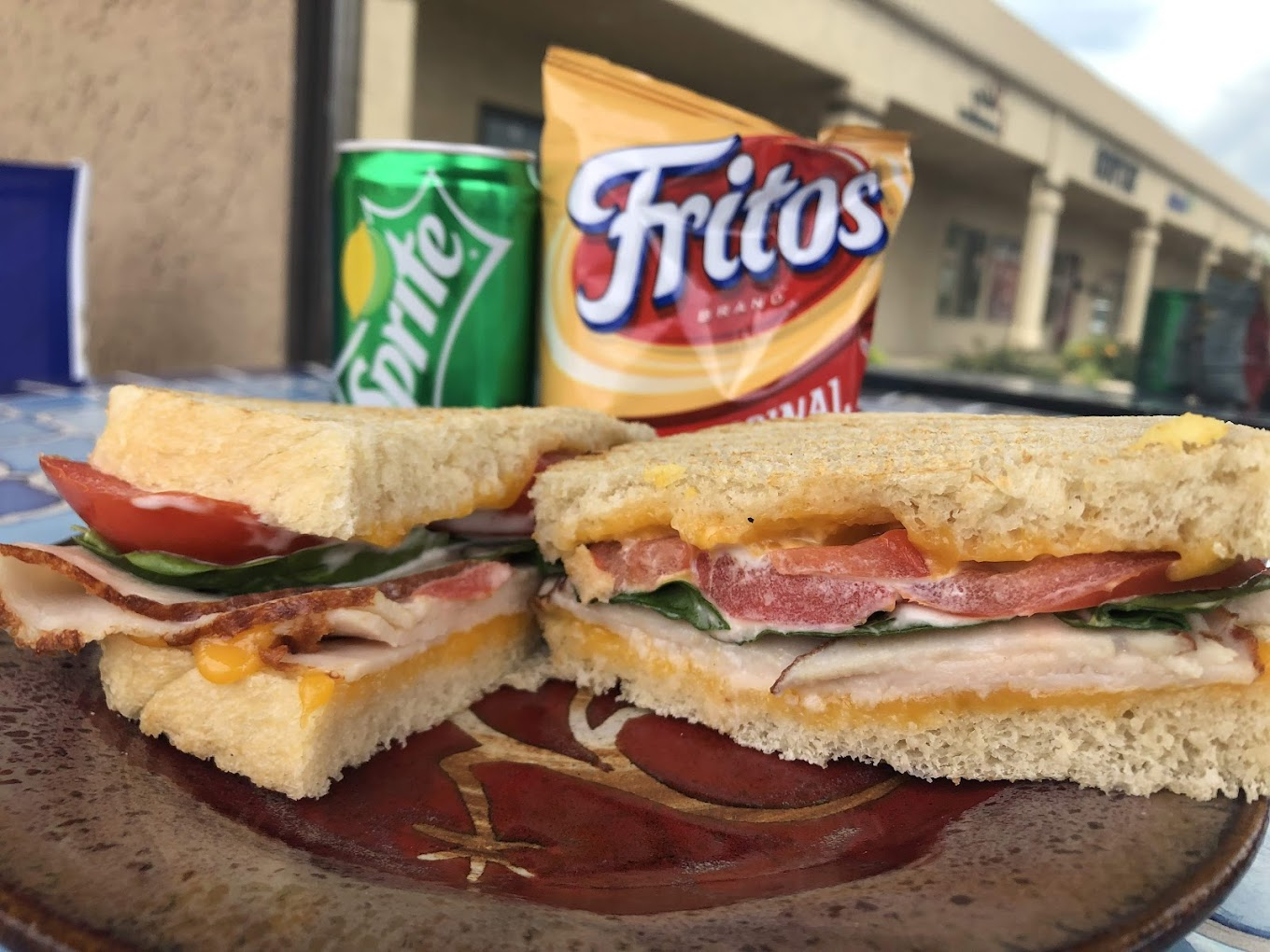 A view of a sandwich cut in half a top of a brown plate, with a side of frito chips and a sprite to drink.