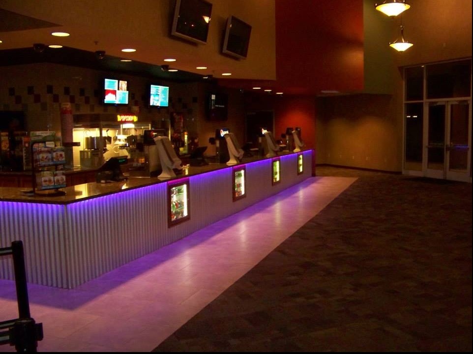 A view of a movie theatre concession stand with LED lights lining the bottom of the counter, TV's displaying the menu line the wall above the counter.