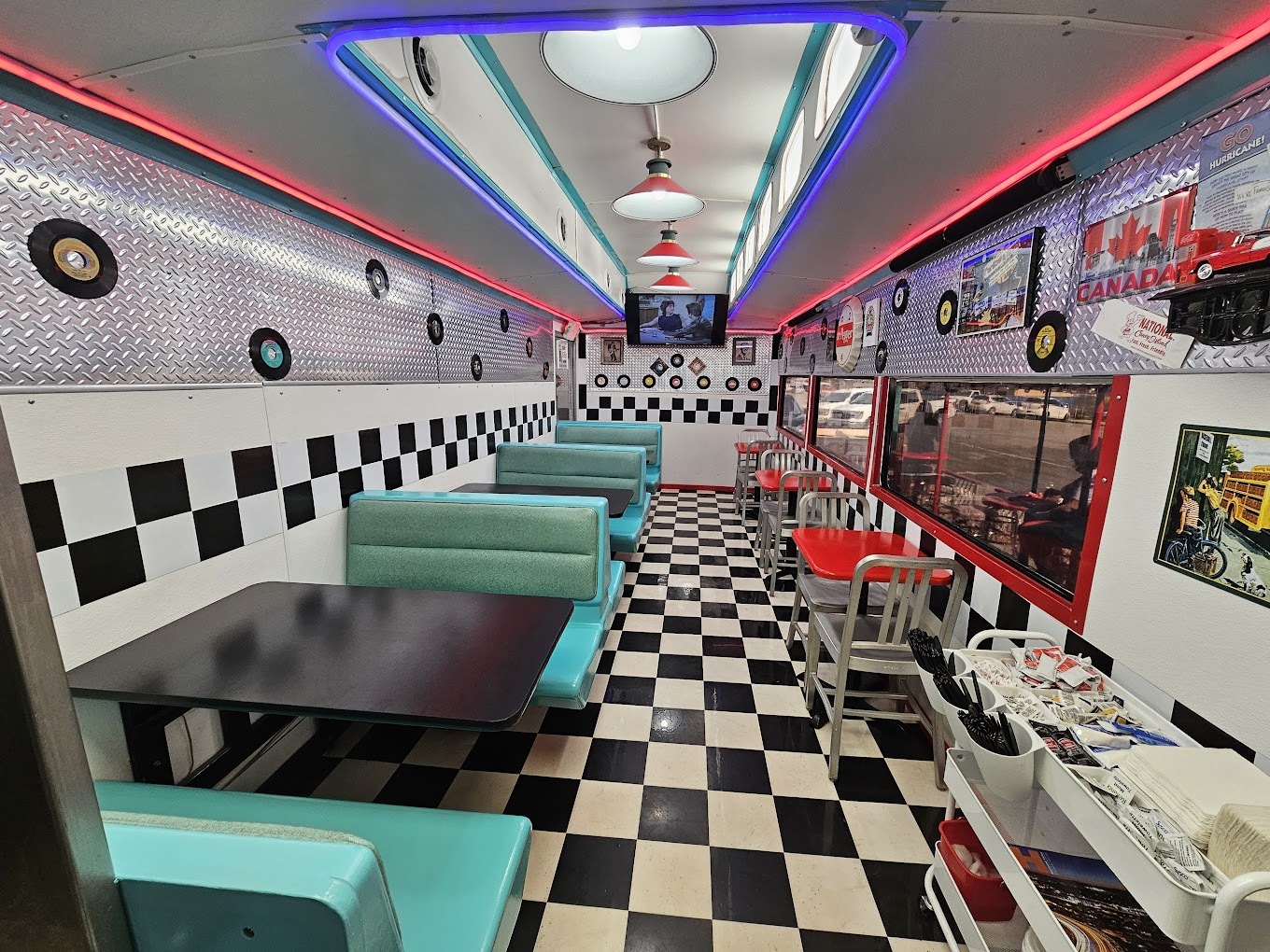 An inside view of a small diner inside of a trailer with checkered flooring and colorful neon lighting.